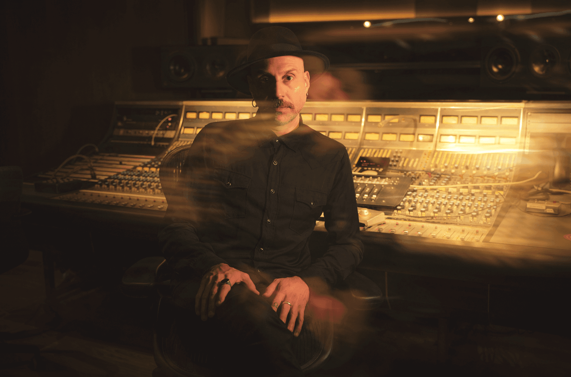 Jaime Gomez-Arellano sitting in a dimly lit recording studio in front of a large analog mixing console, wearing a black shirt and hat.