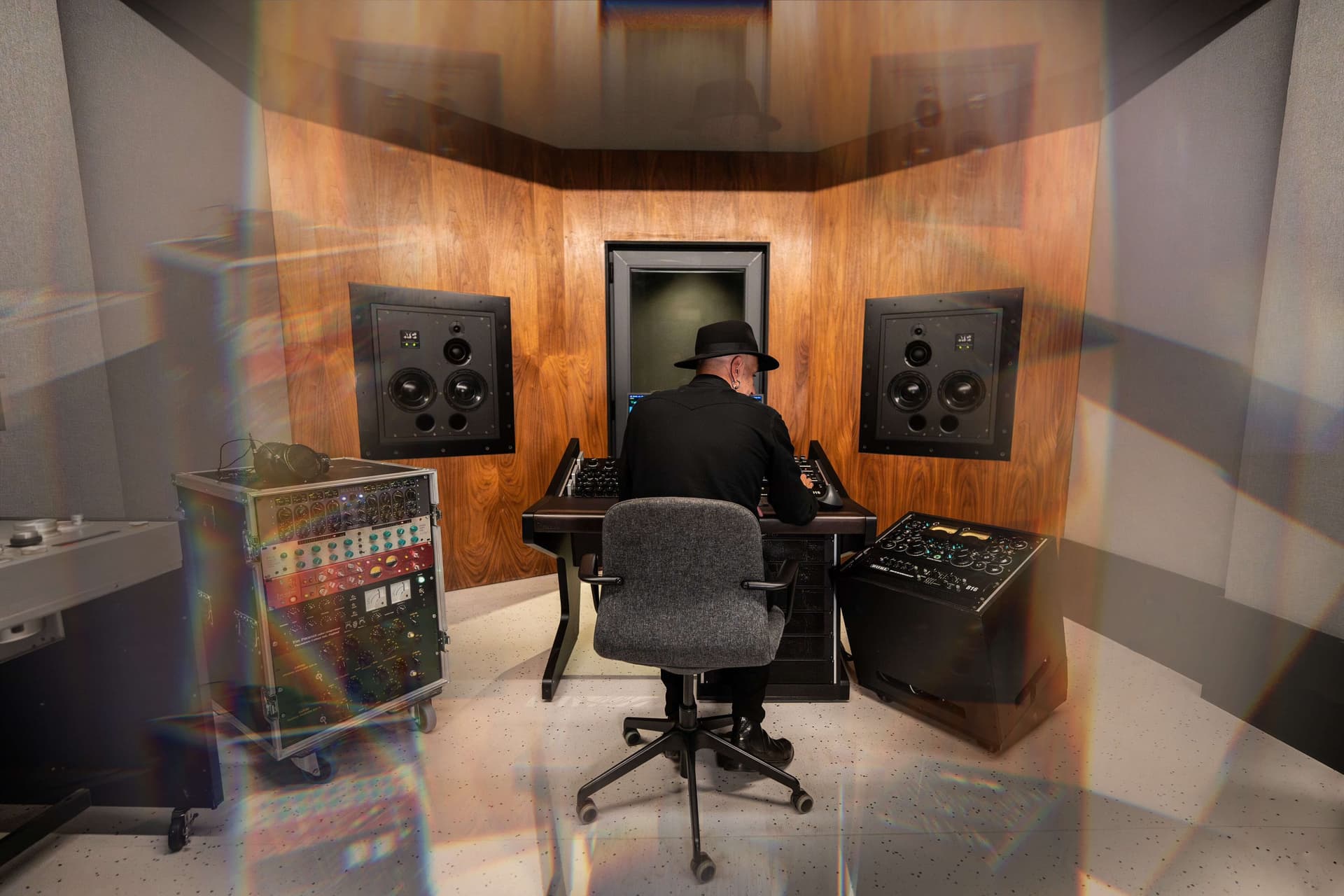 Jaime Gomez-Arellano sitting in a dimly lit recording studio in front of a large analog mixing console, wearing a black shirt and hat.