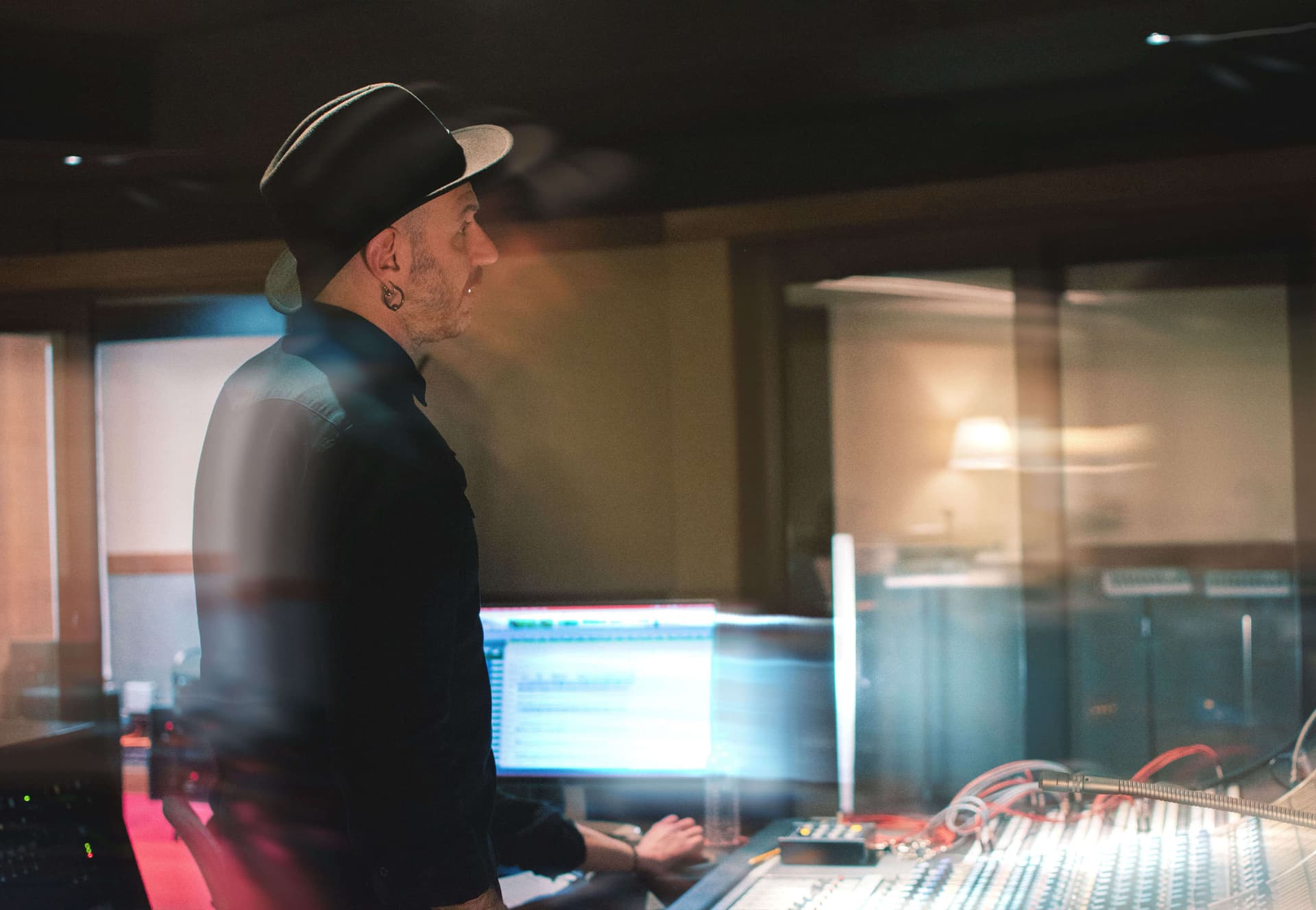 Jaime Gomez-Arellano sitting in a dimly lit recording studio in front of a large analog mixing console, wearing a black shirt and hat.